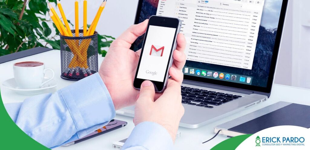 Mano sostiene un móvil con el icono de Gmail frente a un portátil con la bandeja abierta