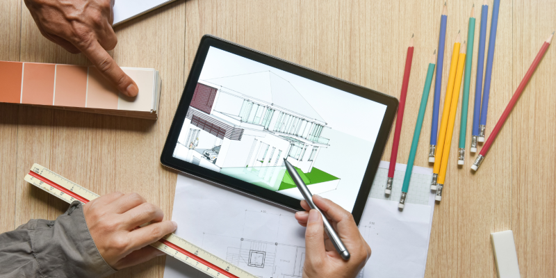 Manos revisan un plano arquitectónico en una tableta junto a lápices de colores y una regla