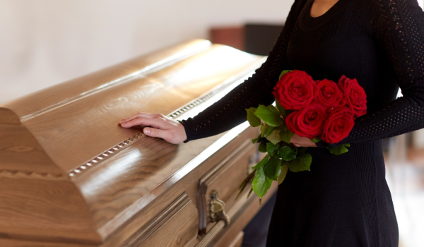 Mujer vestida de negro apoya la mano en un ataúd de madera mientras sostiene rosas rojas
